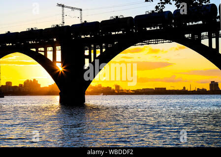 Silhouette einer schönen gewölbten Bahnbrücke und Zug mit Zisternen auf dem Fluss Dnepr bei Sonnenuntergang. Dnipro Stadt, Dnepropetrovsk, Ukraine. Sommer Stockfoto