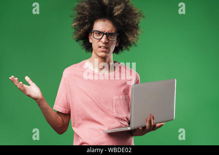 Bild einer jungen verwirrt curly Kerl über grüne Wand mit Laptop Computer Hintergrund posiert. Stockfoto