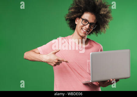 Foto eines Jungen positive lächelnd freundlichen curly Kerl über grüne Wand Hintergrund posiert mit Laptop Computer zeigt auf Copyspace. Stockfoto