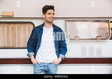 Portrait von glücklichen Mann mit Jeansjacke lächelnd mit den Händen in den Taschen beim Stehen in der Nähe von Haus auf Rädern im Freien Stockfoto