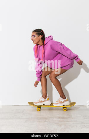 Foto von Wunderschöne emotionale glückliche junge afrikanische Frau skater über weiße Wand Hintergrund in hellem rosa sweatshirt isoliert auf Skateboard. Stockfoto