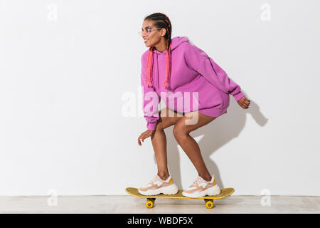 Bild wunderschöne fröhliche junge afrikanische Frau skater über weiße Wand Hintergrund in hellem rosa sweatshirt isoliert auf Skateboard. Stockfoto