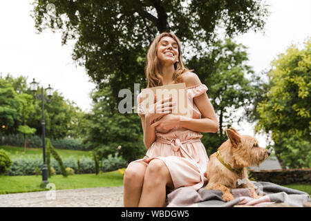 Portrait von Glücklich, aufgeregt Frau in Kleid lesen Buch, während sitzt auf der Bank mit ihrem Hund im Sommer Park Stockfoto