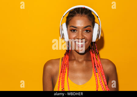 Bild von verlockenden afrikanischen amerikanischen Frau afro Zöpfe lächelnd und Musikhören mit Kopfhörern über Gelb Hintergrund isoliert Stockfoto