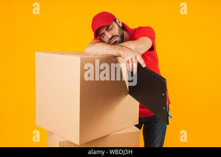 Image der jungen Lieferung Mann in der roten Uniform schlafen im Stehen mit Zwischenablage über Verpackung Kisten über Gelb Hintergrund isoliert Stockfoto