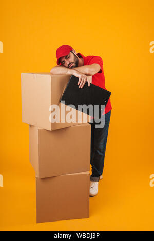 Bild von müde Lieferung Mann in der roten Uniform schlafen im Stehen mit Zwischenablage über Verpackung Kisten über Gelb Hintergrund isoliert Stockfoto