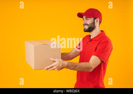 Bild zufrieden Lieferung Mann in der roten Uniform und lächelnd Verpackung über Gelb Hintergrund isoliert Stockfoto