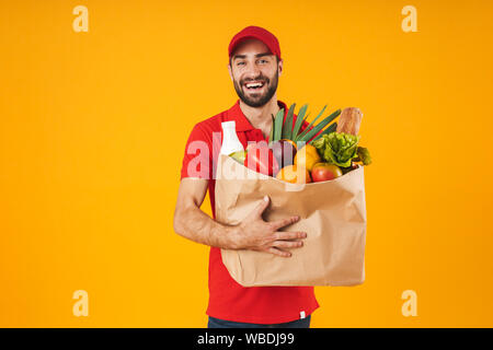 Portrait von zufrieden Lieferung Mann in der roten Uniform Lächeln beim Tragen Papiertüte mit Lebensmitteln über Gelb Hintergrund isoliert Stockfoto