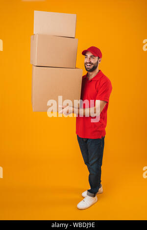 Volle Länge Portrait von zufrieden Lieferung Mann in der roten Uniform Lächeln beim tragen Verpackungskartons isoliert auf gelbem Hintergrund Stockfoto