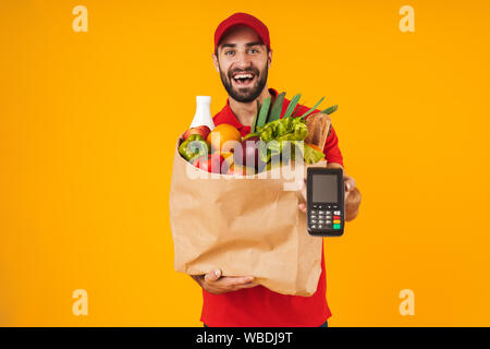 Portrait von zufrieden Lieferung Mann in der roten Uniform holding Payment Terminal und Papier Beutel mit Lebensmitteln über Gelb Hintergrund isoliert Stockfoto