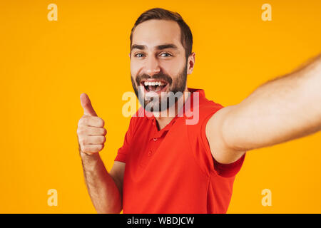 Portrait von erfreut Mann im roten T-Shirt lachen und zeigen Daumen hoch, während der Einnahme von selfie Foto über Gelb Hintergrund isoliert Stockfoto