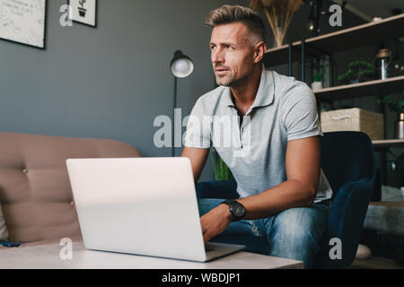 Foto von Konzentriertem kaukasischen Mann in Freizeitkleidung mit Laptop, während auf einem Sessel zu Hause sitzen Stockfoto