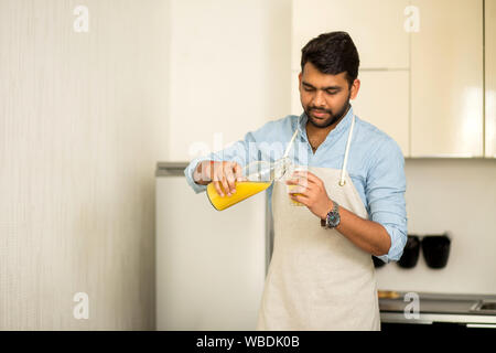 Schön unrasiert indischen Mann mit blauem Hemd und Schürze gießen frische organische Orangensaft in ein Glas an Kamera suchen, prepearing Frühstück in k Stockfoto