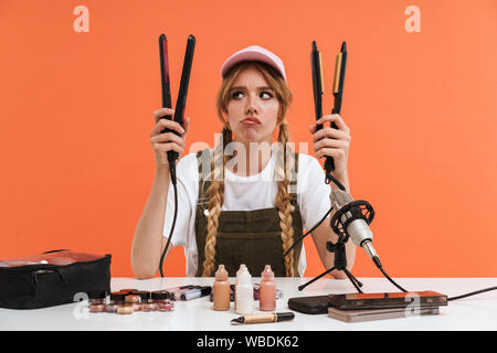 Bild von Sad girl Holding Haarglätter während der Aufnahme blog Sendung über neue kosmetische Produkte über orange Wand isoliert Stockfoto