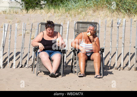 Tayside, Dundee, Schottland, Großbritannien. 26 August, 2019. UK Wetter: Heatwave fegt über Dundee auf Feiertag Montag mit Temperaturen bis 24 Grad Celsius. Urlauber genießen Sie am späten Nachmittag das Wetter auf Broughty Ferry Strand. Zwei Frauen sitzen auf Sitze unten Kühlung Eis essen Kegel. Credit: Dundee Photographics/Alamy leben Nachrichten Stockfoto