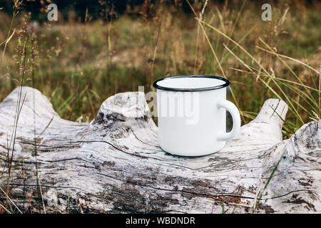 Weißes Metall Becher sitzen, die auf Holz anmelden. Gras, Wiese Hintergrund verschwommen. Outdoor Tee, Kaffee, Mockup von Emaille Tasse. Lifestyle entspannen, Trekking Stockfoto