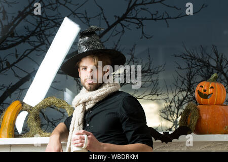 Halloween Mann, der Hexe Hut und Schal. Zaubertrank Konzept. Macho holding Schale am Fenster mit Bäume im Herbst. Urlaub Symbole und Dekorationen. Warm Stockfoto