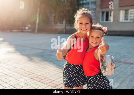 Glückliche Schwestern Mädchen, Rucksäcke und Daumen hoch. Kinder Schüler lächelnd im Freien Schule. Bildung. Zurück zur Schule Stockfoto