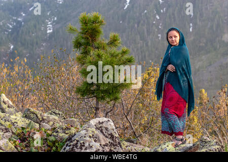 Ein nettes Mädchen in einem Kleid und Schal steht auf großen Steinen neben einem jungen Nadelbaum, in den Bergen. Vor dem Hintergrund der Berge mit Con abgedeckt Stockfoto