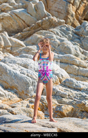 Süße kleine Mädchen gegen Rocky Hintergrund mit Händen wie Sonnenbrille posieren, in voller Badeanzug Stockfoto