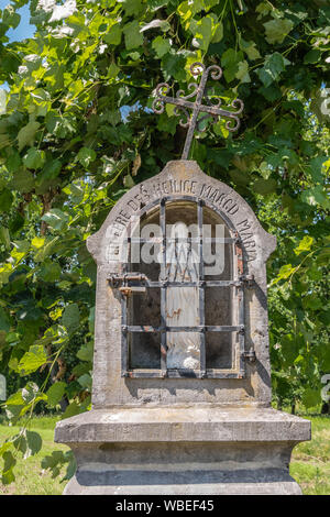 Bokrijk, Belgien - 27 Juni, 2019: Nahaufnahme von kleinen grauen Stein Kapelle Nische entlang der Straße mit weißen Statue der Jungfrau Maria. Iron Cross auf die Oberseite ist schräg. Stockfoto