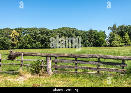 Bokrijk, Belgien - 27 Juni, 2019: Nahaufnahme von alten grauen hölzerne Tor Verschluss mit Baum Schwebebalken, um das Öffnen zu erleichtern. Grüne Wand von Bäumen unter blau Stockfoto