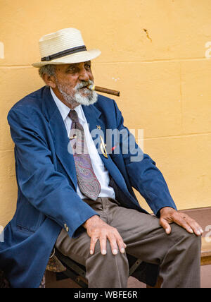 Trinidad, Kuba, 26. November 2017 - Der alte Mann sitzt auf der Bank das Rauchen einer Zigarre, gegen eine gelbe Wand Stockfoto
