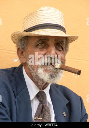 Trinidad, Kuba, 26. November 2017 - Der alte Mann sitzt auf der Bank das Rauchen einer Zigarre, gegen eine gelbe Wand Stockfoto