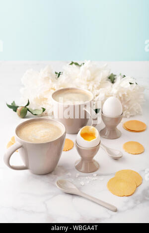 Weich gekochte Eier mit liquide orange Eigelb in Keramik Eierbecher, zwei Tassen Kaffee mit keramischen Löffel und dünne knusprige Mais Chips auf dem Hintergrund der Beau Stockfoto