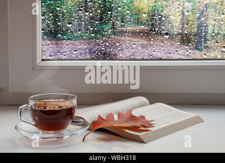 Tasse Kaffee, eine offene Bibel und einem roten Ahornblatt auf dem Fensterbrett mit Regentropfen auf das Glas auf düsteren Herbst Tag Stockfoto