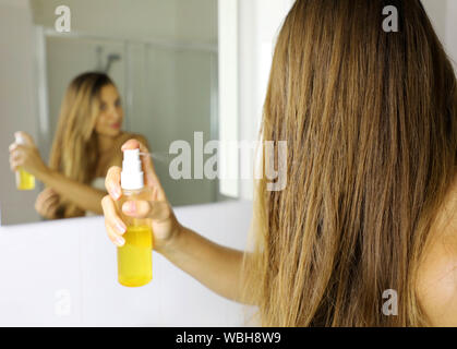 Junge Frau mit unter Druck stehendem Öl Maske Spray auf Haar Tipps vor einem Spiegel. Haarpflege Konzept. Fokus auf das Haar. Stockfoto