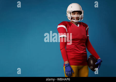 Bild des amerikanischen weibliche Fußball-Spieler in weißen Helm, Sportswear und Kugel in ihre Hand an den leeren blauen Hintergrund. Für text Stockfoto