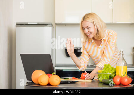 Freudige blonde Frau Abendessen kochen und mit Laptop in der Küche, in video. Glücklich lächelnde Frau mit Freund mit Anwendung auf Comp Stockfoto