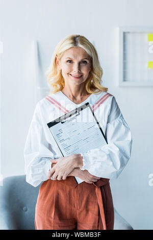 Fröhliche blonde Frau mit Zwischenablage mit Lebenslauf im Büro Stockfoto