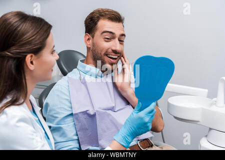 Selektiver Fokus der bärtige Patienten berühren Gesicht halten Spiegel in der Nähe Zahnarzt Stockfoto