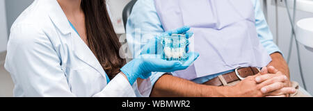 Panorama-aufnahme des Zahnarztes in Latex Handschuhe holding Zähne Modell in der Nähe von Patienten mit geballten Händen Stockfoto