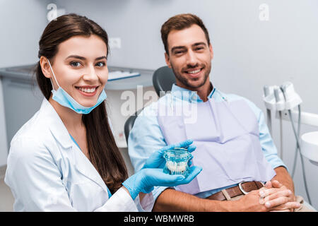 Selektiver Fokus von attraktiven Zahnarzt in Latex Handschuhe holding Zähne Modell in der Nähe von Patienten mit geballten Händen Stockfoto