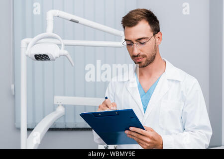 Schöner Mann im weißen Kittel und Brille, Stift und Zwischenablage in der zahnmedizinischen Klinik Stockfoto