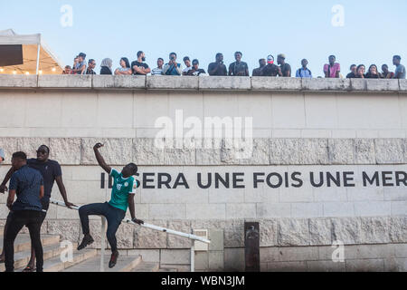 LYON, Frankreich - 14. JULI 2019: Junge schwarze Französische Mann sitzt auf der Treppe eine selfie mit seinem Smartphone vor einer Masse am Ufer des Stockfoto