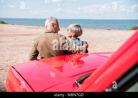 Rückansicht des Senior Paar in der Nähe von roten Auto am Strand und in sonnigen Tag Stockfoto