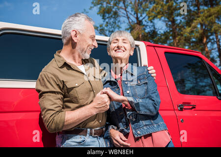 Lächelnd senior Paar Touristen in der Nähe von roten Auto, halten sich an den Händen und im sonnigen Tag Stockfoto