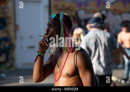 Ein schwarzer Mann rauchen im Notting Hill Carnival trägt ein rosa Halsband, reflektierend Sonnenbrille und eine bunte Perücke. Stockfoto