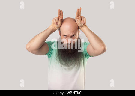 Porträt von Ernst oder böse Glatze im mittleren Alter Mann mit langem Bart in grünem t-shirt mit Finger Hörner auf dem Kopf und Suchen mit aggressiven Gesicht Stockfoto