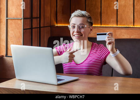 Portrait von schöne glückliche junge bussineswoman mit kurzen Haaren in Rosa t-shirt und Brillen im Cafe sitzt, Zeigefinger zu Kreditkarte, Eas Stockfoto