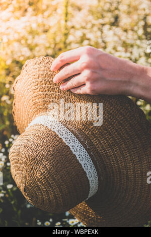 Halten Sie einen Strohhut in warmen Nachmittag Sonne. Weibliche hand mit einem Hut auf ein wunderschönes ländlichen Wiese voller wilder Blumen, vintage Film Stockfoto