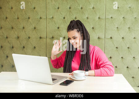 Hallo. Sympathische junge Frau mit schwarzen dreadlocks Frisur in rosa Bluse sitzt und Gruß Arbeitnehmer durch eine Webcam zum Laptop und winkte ihr Stockfoto