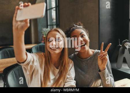 Porträt von zwei Multirassischen junge Frauen, die selfies mit Smartphone, wie sie in der Cafeteria sitzen. Horizontale erschossen. Ansicht von vorn Stockfoto