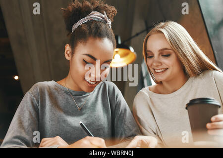 Nahaufnahme, Porträt von zwei Multirassischen junge weibliche Freunde diskutieren neue Ideen und Notizen in ein Notebook, wie sie in der Cafeteria genießen eine Tasse Kaffee sitzen. Horizontale erschossen. Ansicht von vorn Stockfoto