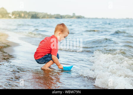 Happy kaukasischen Rothaarige Kleinkind Kind Junge mit einem blauen Papier Boot im Wasser auf See Meer Meer bei Sonnenuntergang am Abend oder Morgen. Kid spielen auf Bea