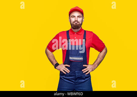 Porträt der jungen zuversichtlich Handwerker mit Bart in Blau, Rot T-Shirt und Cap stehen und halten sich an den Händen an der Hüfte mit einem Lächeln, Innen, Studio s Stockfoto